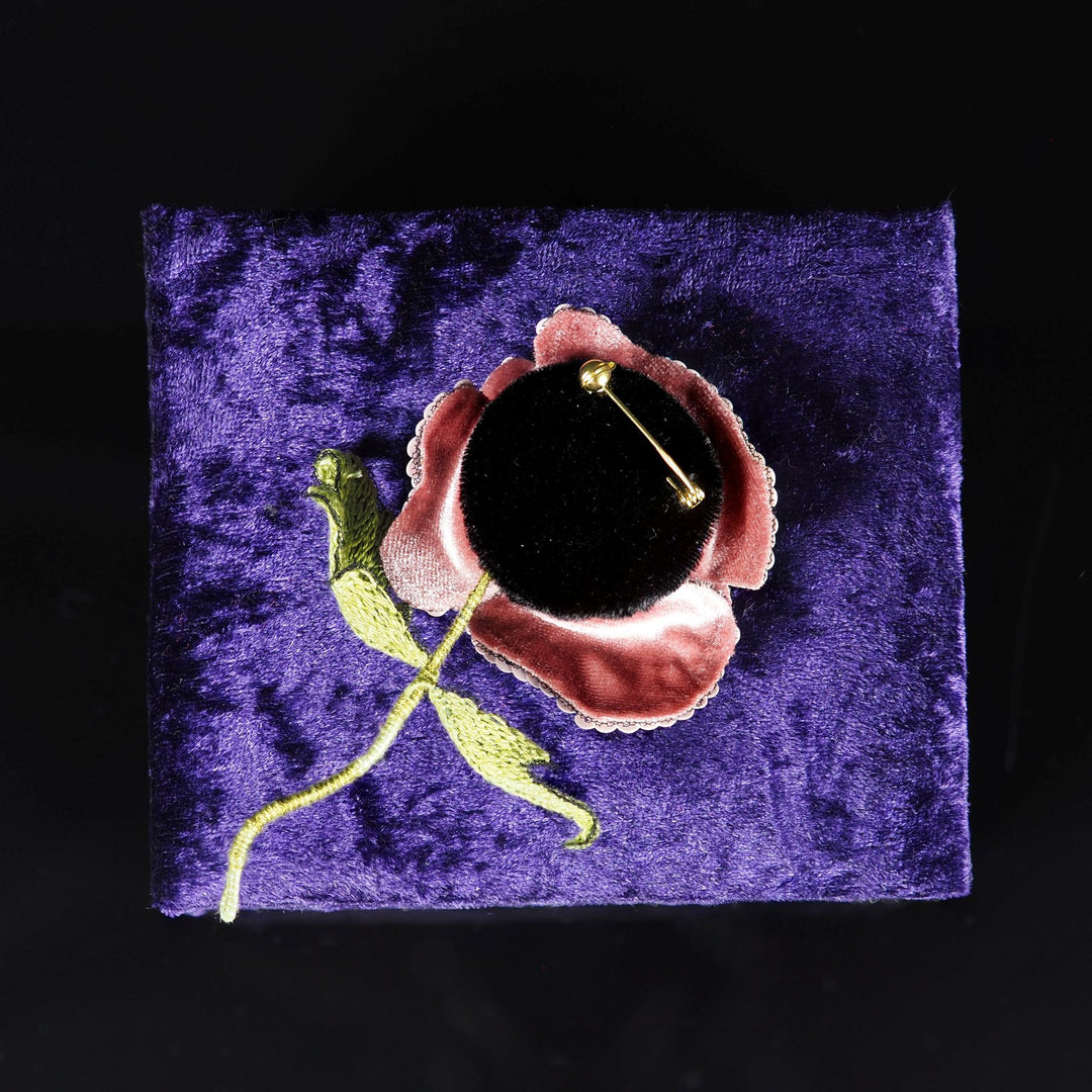 Back side of blush pink poppy brooch showing velvet-covered base and secure gold-tone pin with safety ball, displayed on a purple velvet stand
