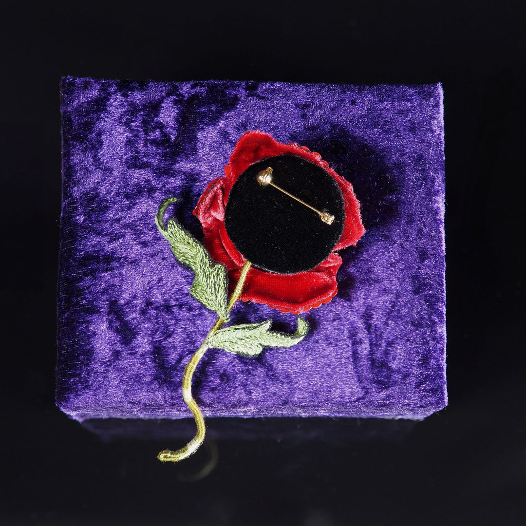 Back side of handmade red poppy brooch showing black velvet backing and secure pin with safety ball, displayed on a purple velvet stand