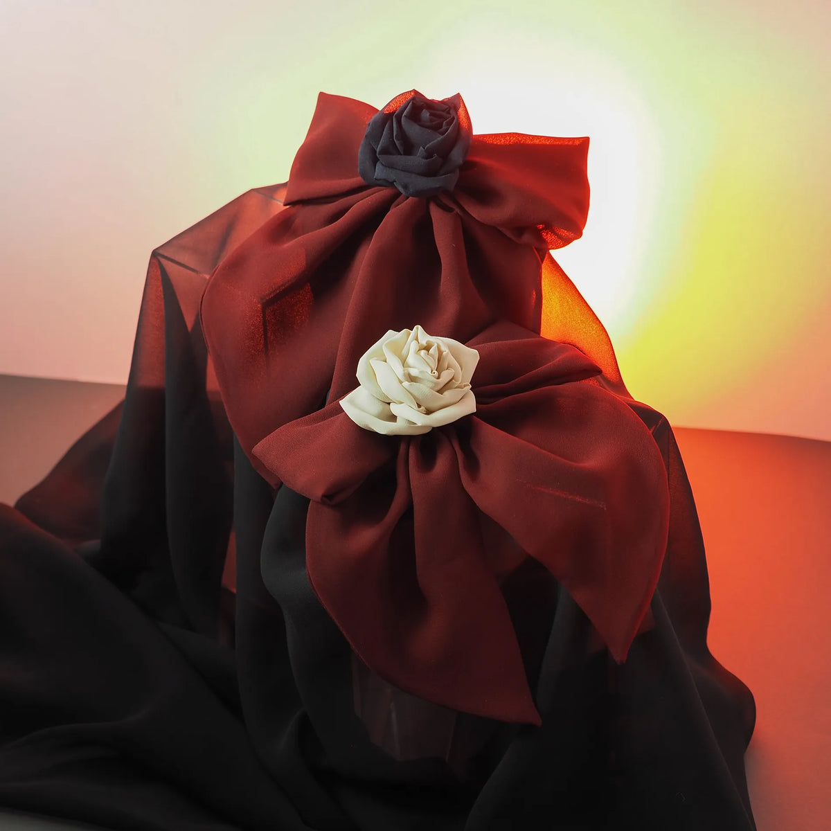 Two handmade silk rose hair clips, one with a black rose and one with an ivory rose, arranged on large red chiffon bows and styled on sheer black and red fabric with warm lighting. Shadow & Bloom collection by BY ROVA