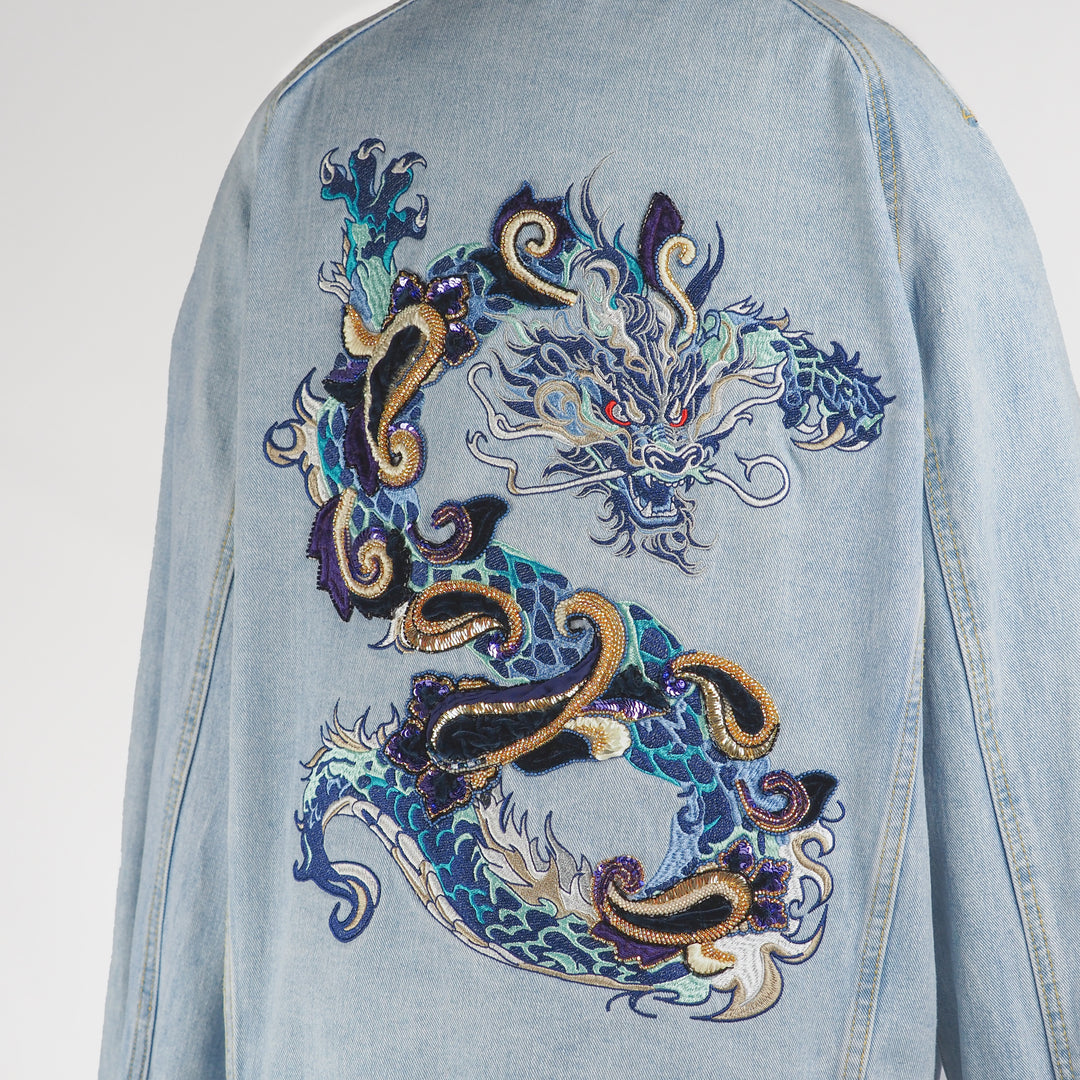 Close-up of embroidered dragon design on the back of an oversized light blue denim jacket, showing detailed stitching in blue, gold, white, and black threads with beaded accents.