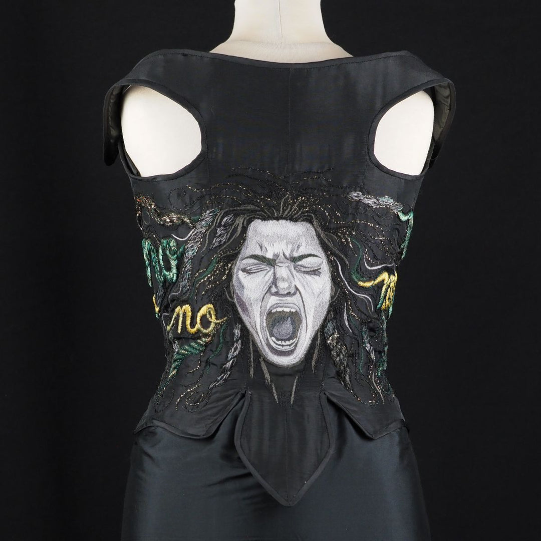 Black taffeta corset with hand-embroidered Medusa face and rope-like hair forming the word “NO” across the back, displayed on mannequin.