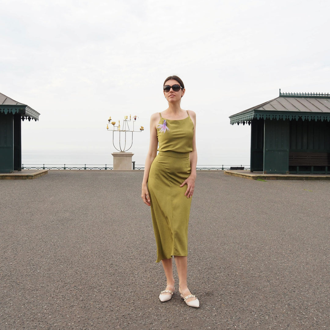 Full-body image of a woman wearing a green dress with a handmade purple hummingbird brooch, photographed outdoors on the British coast. Unique textile accessory designed in the UK