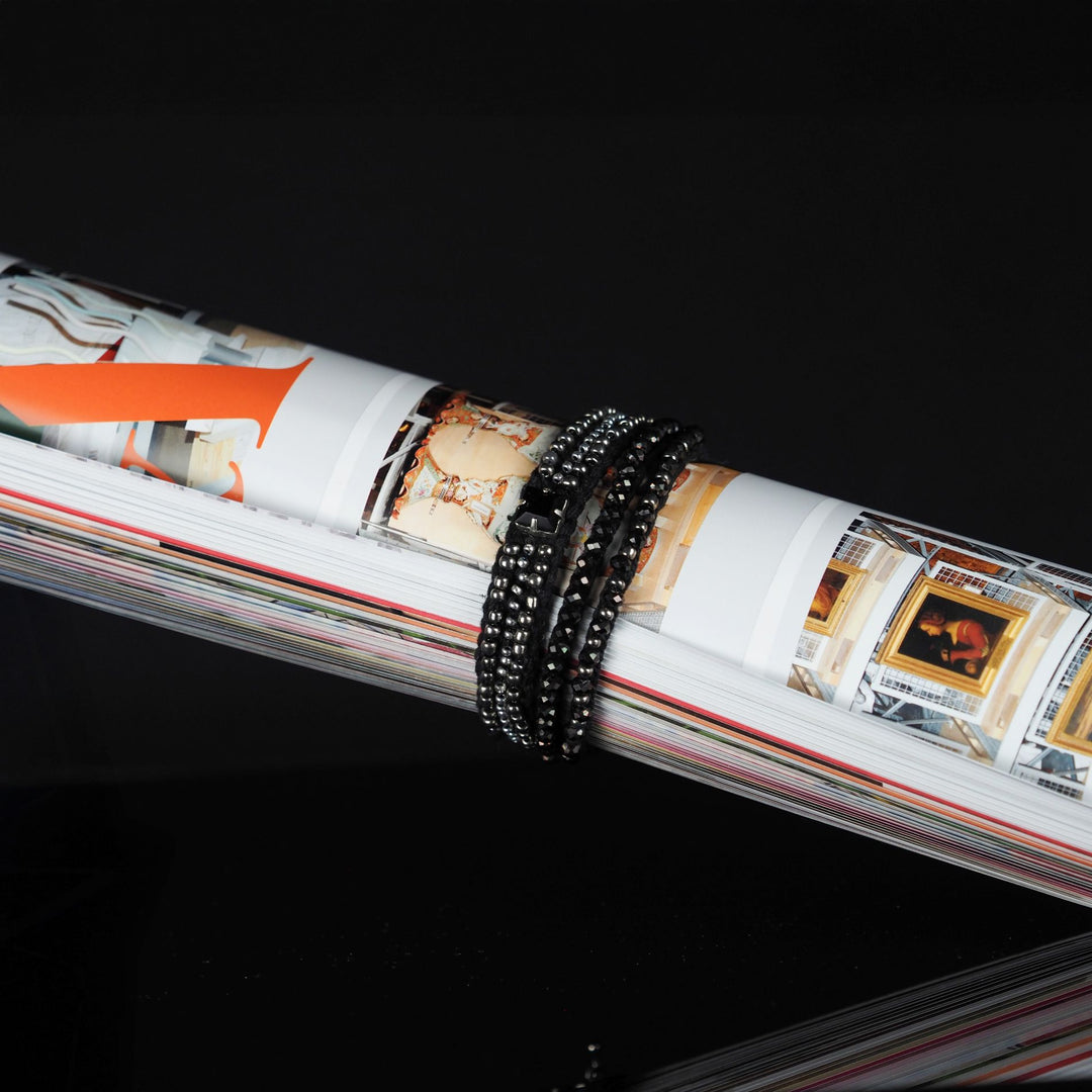 Black and silver multi-strand beaded bracelet displayed wrapped around an open art magazine, handmade on a textile base with glass beads and rhinestone detail.
