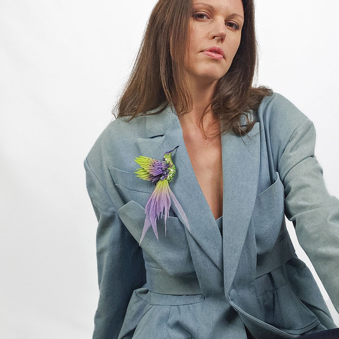Woman wearing a light blue jacket with a lime and purple hummingbird brooch pinned to the lapel, showing scale and styling of the accessory.