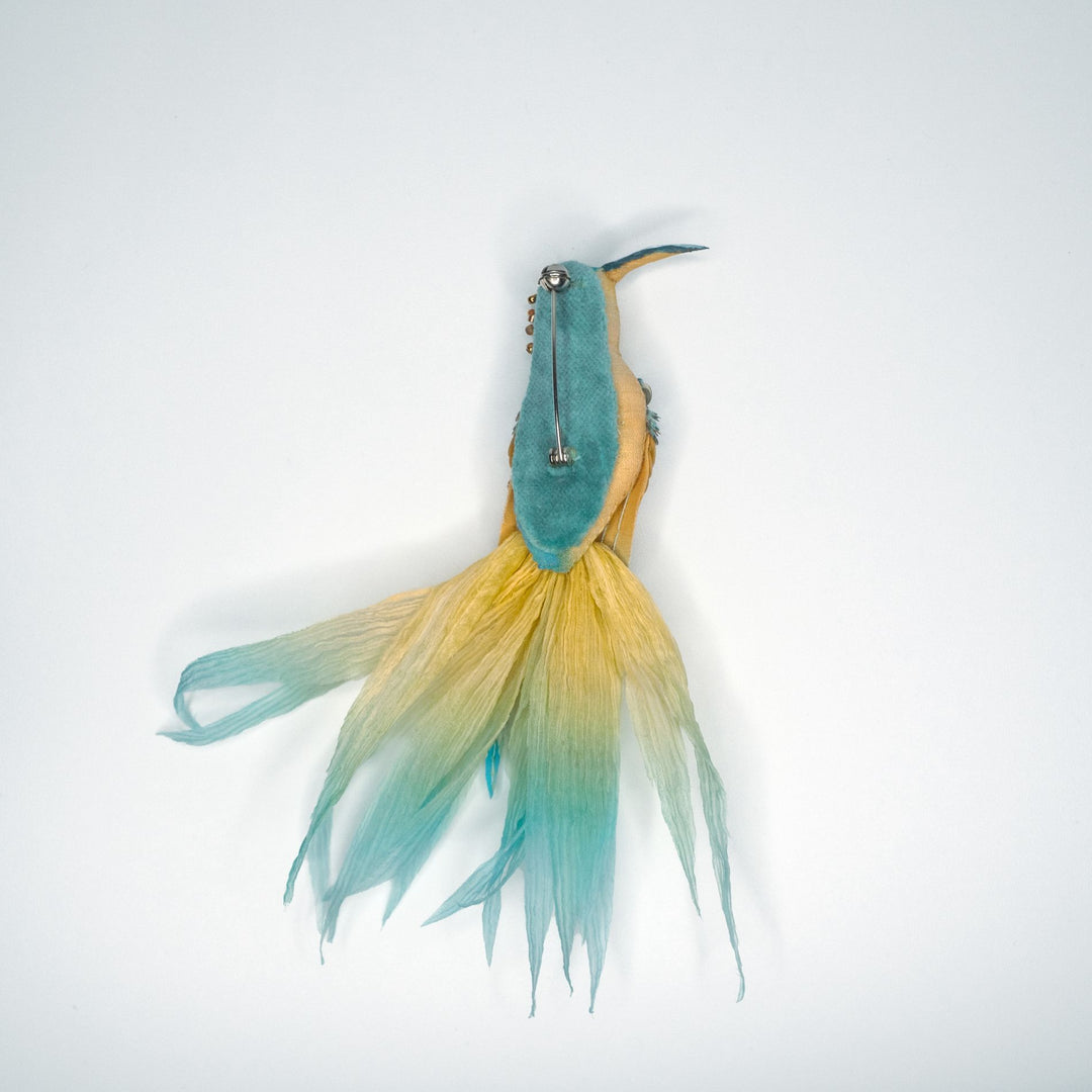 Back view of handmade hummingbird brooch pin showing teal velvet backing, metal clasp, and flowing golden teal feathered tail