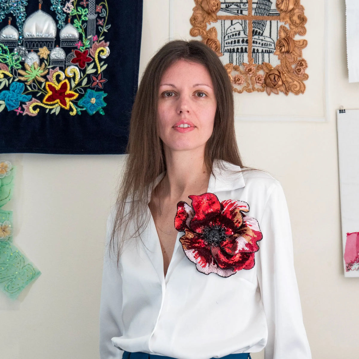 Portrait of Ksenia Semirova, hand embroidery and textile artist, wearing a white blouse with a large red sequined floral brooch, standing in front of embroidered artworks in her studio.