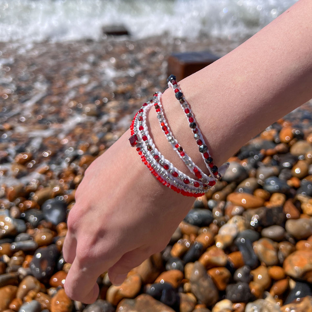 Close-up of red and white multi-strand beaded bracelet on wrist against pebbled beach and ocean waves, handmade boho jewellery.