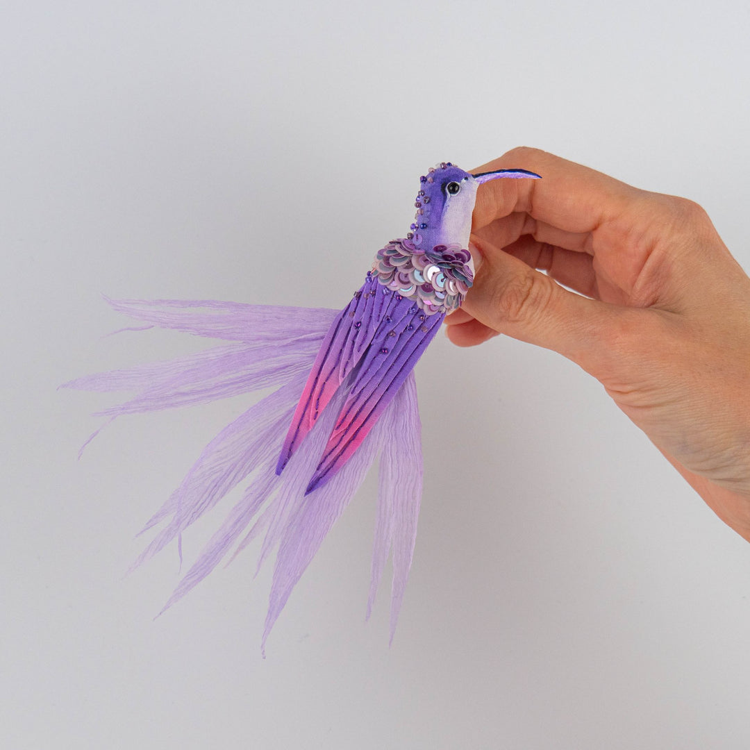 Hand holding purple and white embroidered hummingbird brooch, showing size of unique textile accessory, handmade in the UK