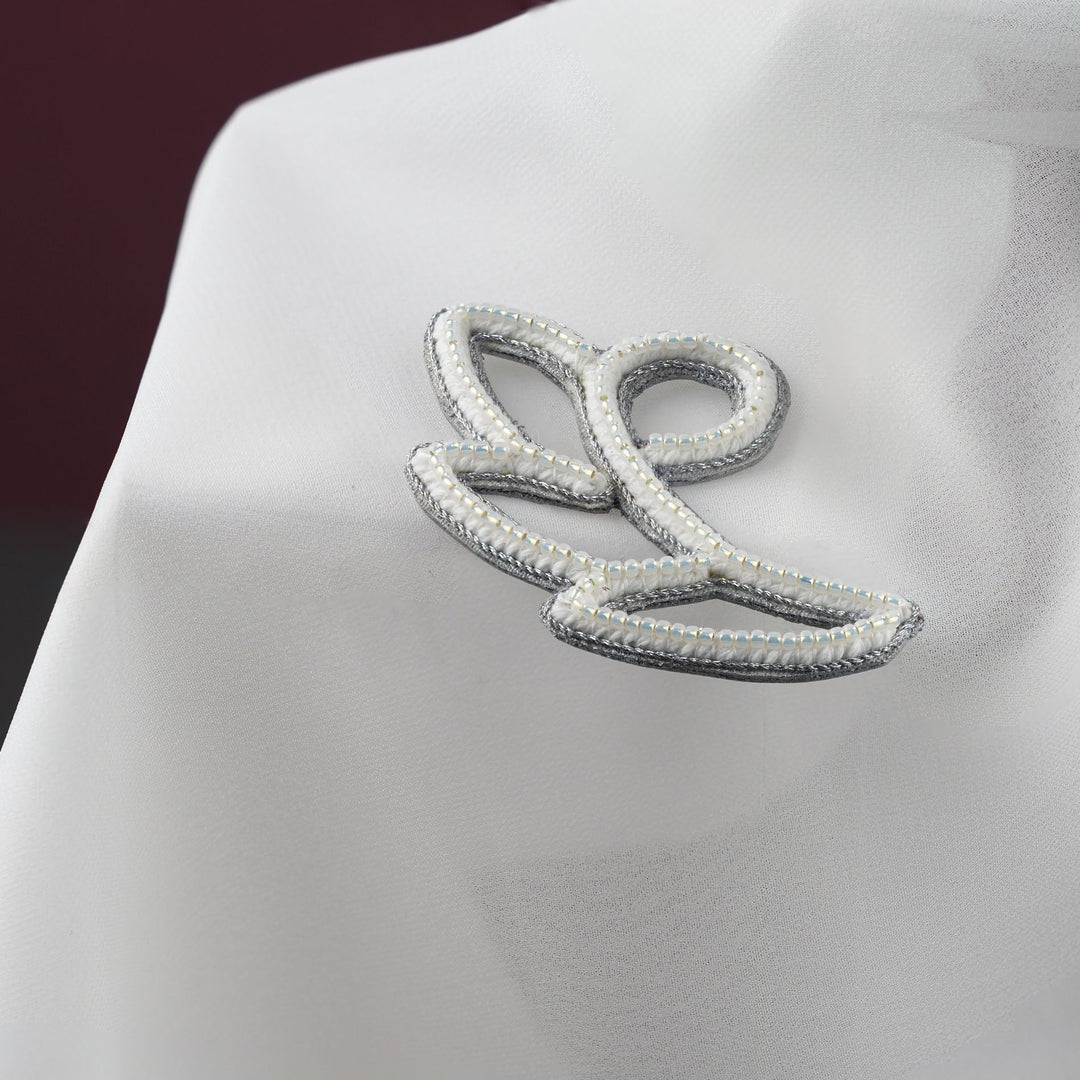 Close-up of handmade white and silver floral bag charm, with seed beads and metallic cord detail, shown on white fabric