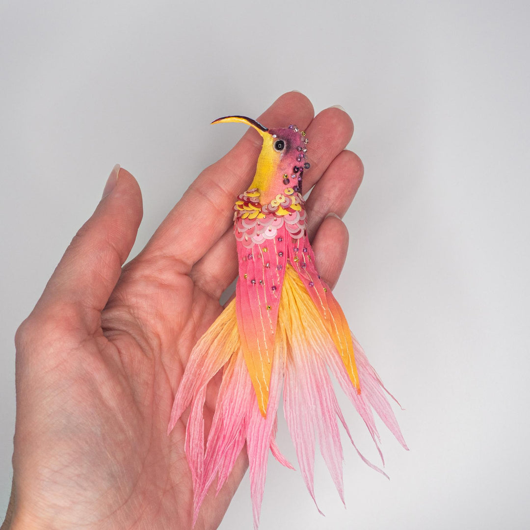 Small yellow and pink hummingbird brooch shown in hand to illustrate size and detailed craftsmanship.