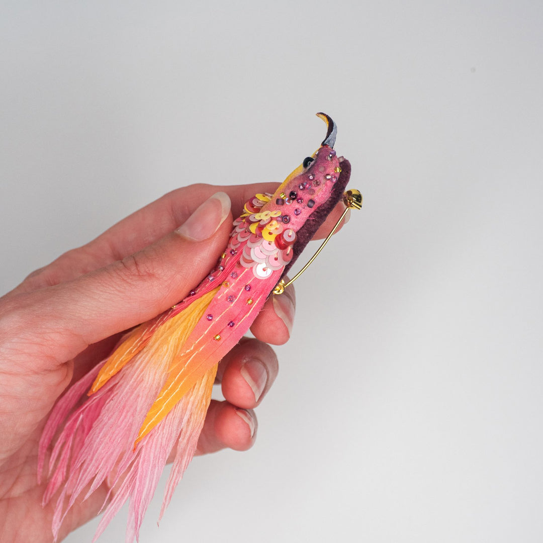 Side profile of the hummingbird brooch in a hand, showing layered sequins and stitched fabric tail.
