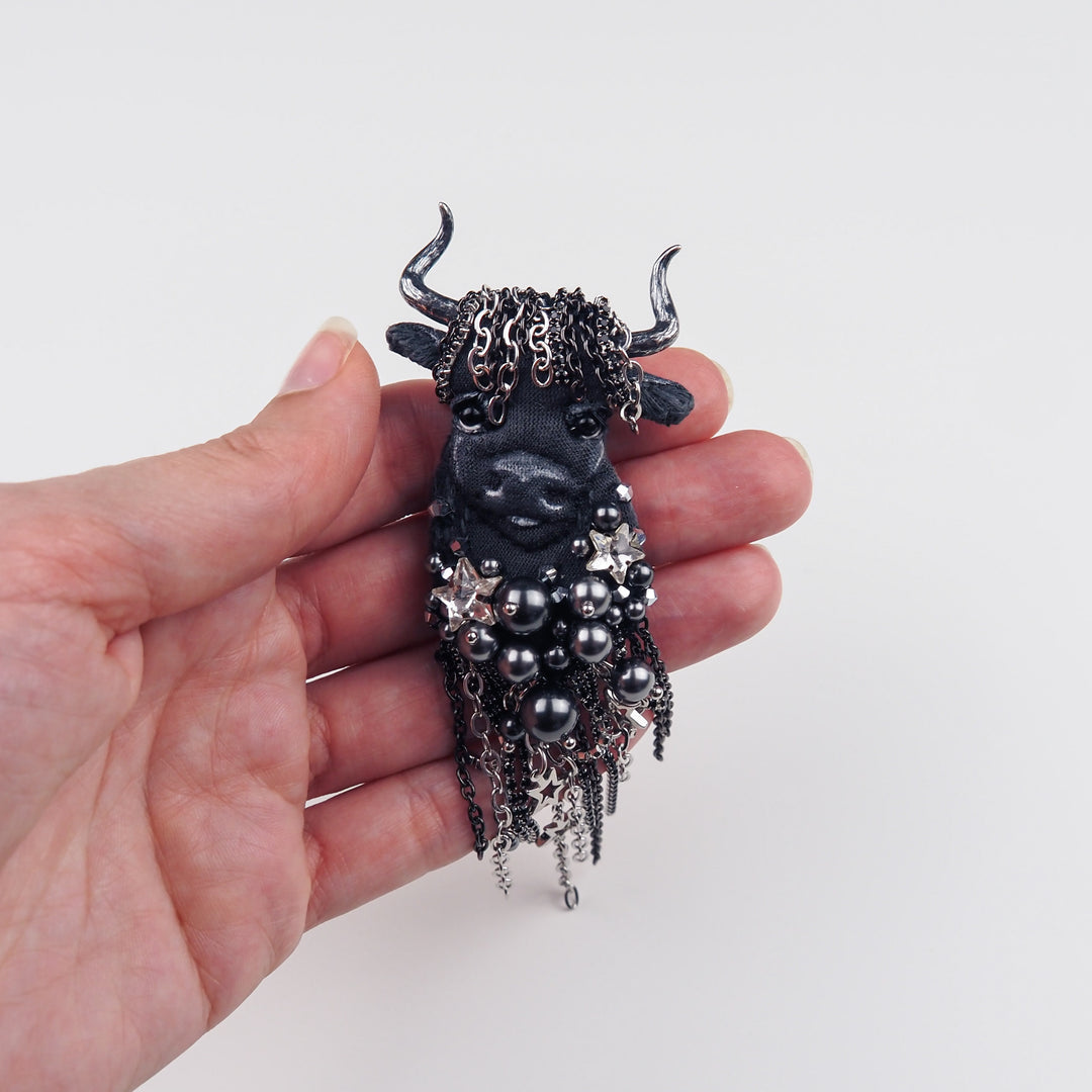Close-up of Taurus brooch held in hand to show size and details of embroidery, custom horns, pearls, and chain fringe.