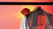 Close-up of an red silk rose choker with black ties draped over a white blouse on a hanger, photographed against a soft red-orange background