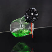 Handcrafted black dahlia flower brooch with pearl centre and red bead drops, styled with green liquid in a glass for a Dark Romance Halloween theme