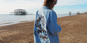 Woman wearing an oversized denim jacket with hand-embroidered dragon motif, standing on Brighton beach near the West Pier. Banner image for embroidered dragon denim jacket product by BY ROVA