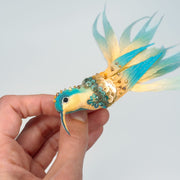 Handmade teal and gold hummingbird brooch pin shown in hand, highlighting embroidery, sequins, and long feathered tail.