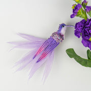 Handmade purple hummingbird brooch with feathered details, styled with fresh purple flowers on a white background. Designed by BY ROVA