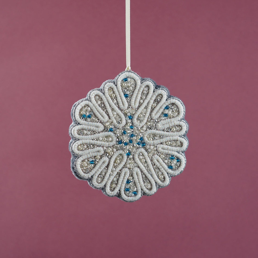 Hanging view of the Jewels of Winter circular snowflake ornament by Ksenia Semirova, crafted with pearl-setting technique in white, silver, and blue tones. Shown suspended on a satin ribbon against a pink background.