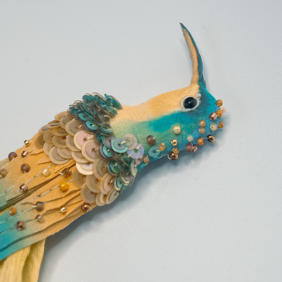 Detailed close-up of hummingbird brooch head, featuring teal and golden fabric with hand-embroidered sequins and beads.