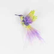Hand-embroidered hummingbird brooch in lime green and purple with sequins, beads, and a long ombré textile tail, photographed on a white background.