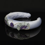 Side view of a lavender satin padded headband with hand-stitched poppy motifs in purple sequins and grey chiffon, decorated with rhinestone chain, resting on a black reflective surface