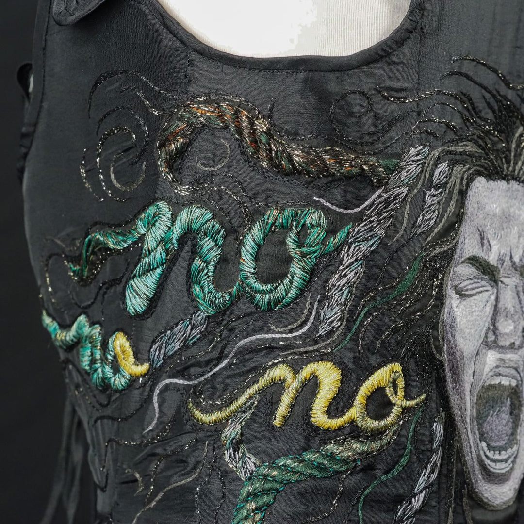 Detailed view of hand embroidery on black corset, showing textured tambour and padded stitches forming Medusa’s hair and stitched word “NO.”