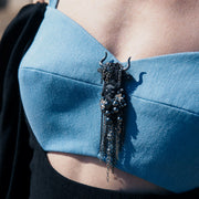 Taurus textile brooch with black embroidery and chain fringe, worn on a blue structured top as a statement accessory